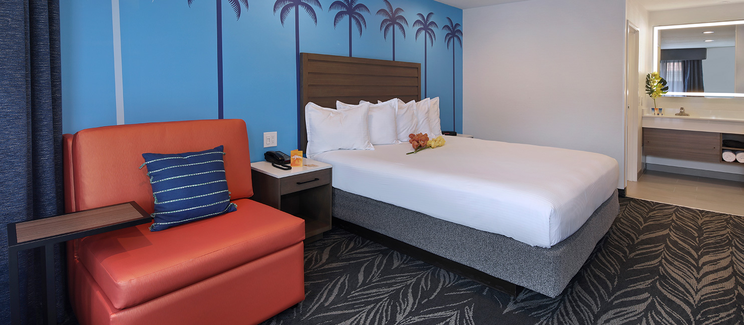 A hotel room with a bed, chair, nightstand, and bathroom in the background.