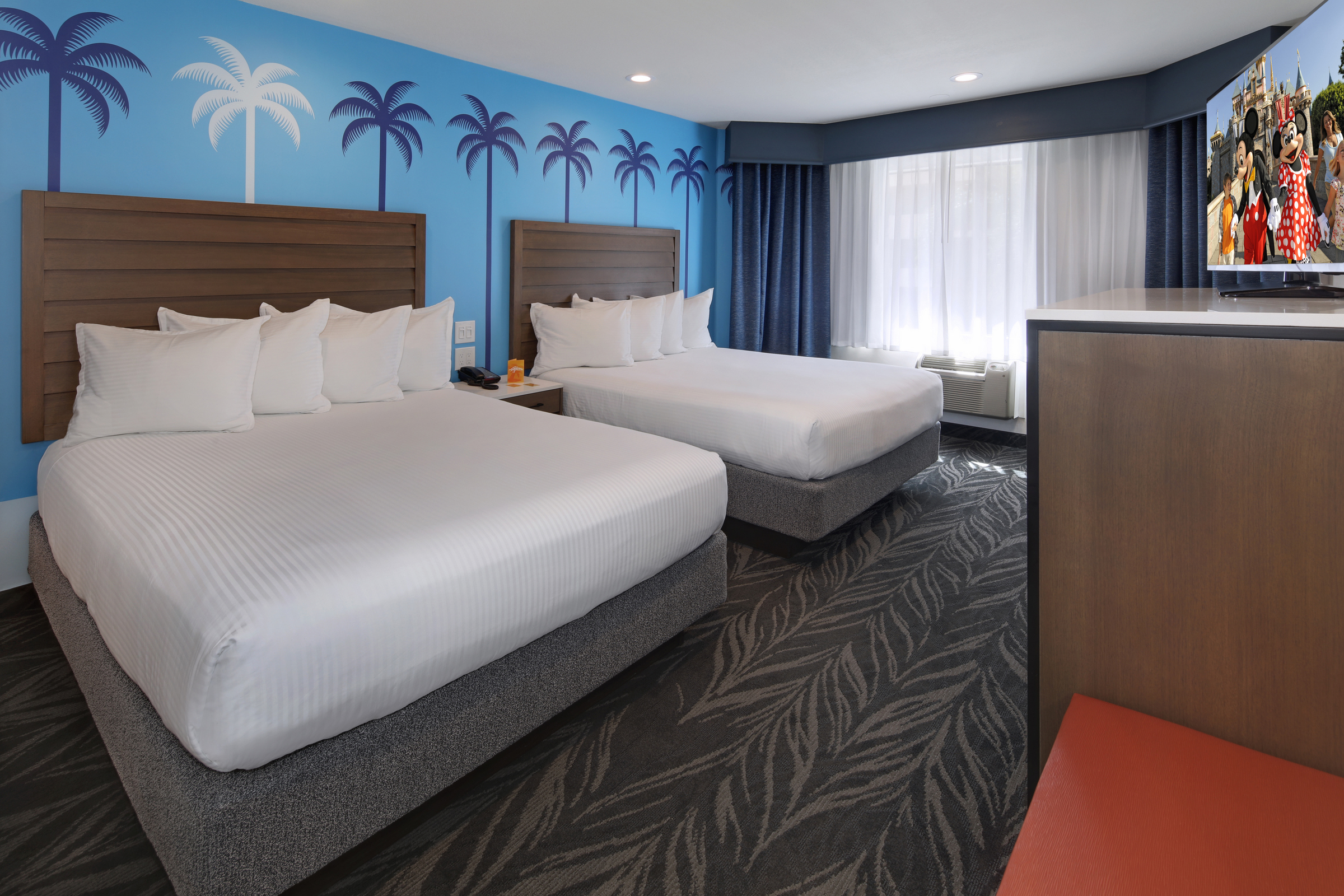 A hotel room with two queen beds, a flat-screen TV, and a window with palm tree wallpaper.