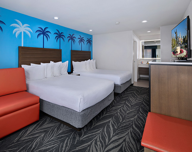 A hotel room with two queen beds, a red sofa, a flat-screen TV, and a kitchenette, featuring palm tree wallpaper.