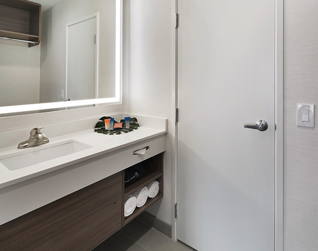 A modern bathroom sink area with a large mirror, a white countertop, and a wooden cabinet.