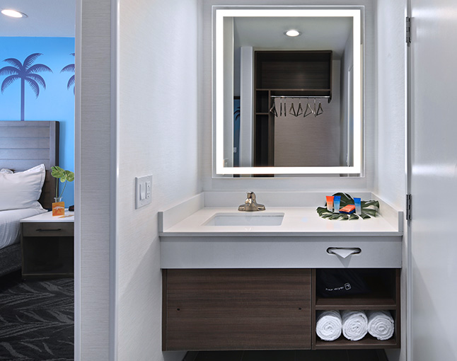 A bathroom sink area with a large mirror, towels, and a view of a bed in the background.