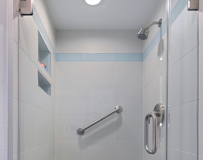 A shower stall with white tiles, a blue accent stripe, a grab bar, and a glass door.