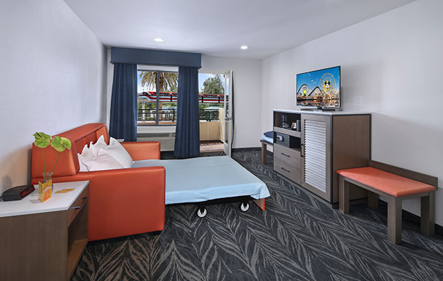 A hotel room with a sofa bed, desk, TV, and balcony