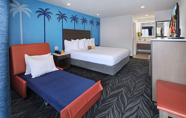 A hotel room with a queen bed, pull-out sofa bed, and palm tree wallpaper.