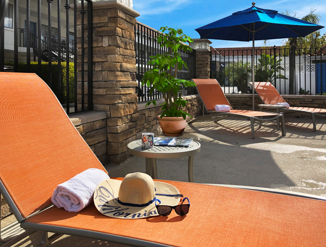 A poolside scene with orange lounge chairs, a towel, hat, sunglasses, and an umbrella.