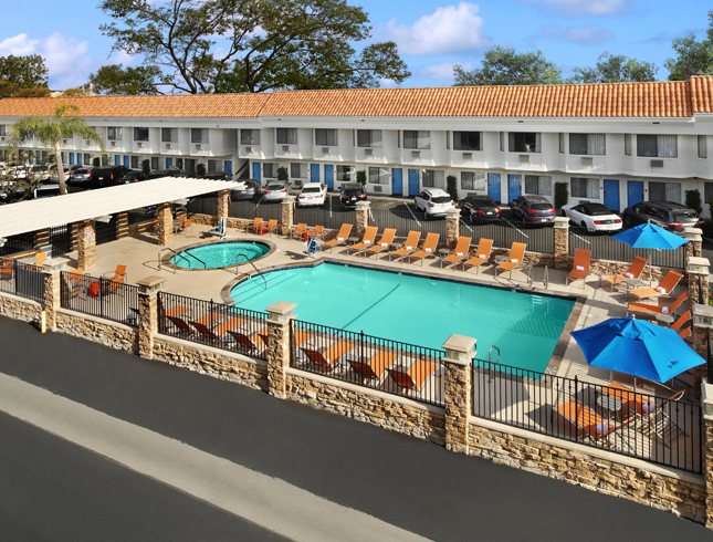 A hotel pool area with a large pool, jacuzzi, lounge chairs, and umbrellas in front of a two-story hotel building.