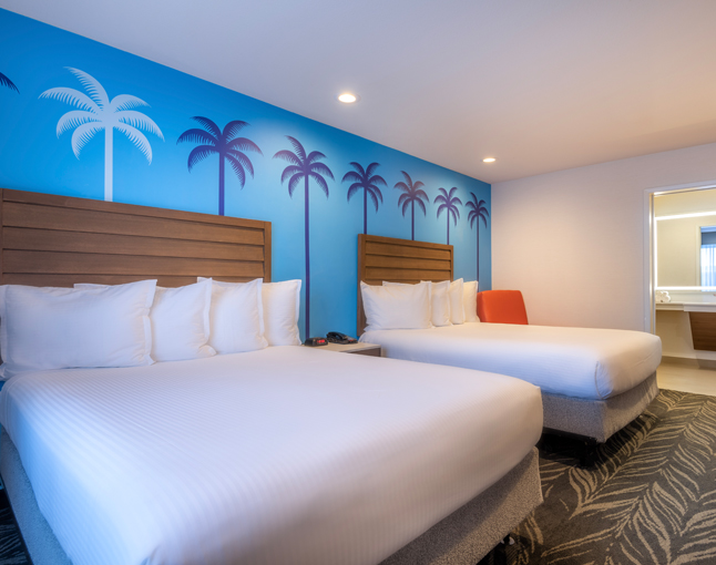 A hotel room with two queen beds, a blue wall with palm tree murals, and a partially open bathroom door.