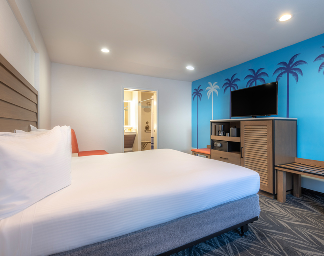 A hotel room with a large bed, TV, and palm tree mural